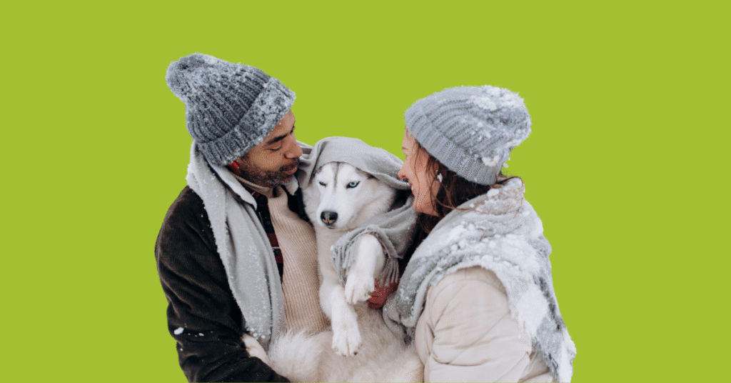 Happy couple holding a dog wearing warm winter clothes outside in the snow, illustrating how to keep pets safe this winter.